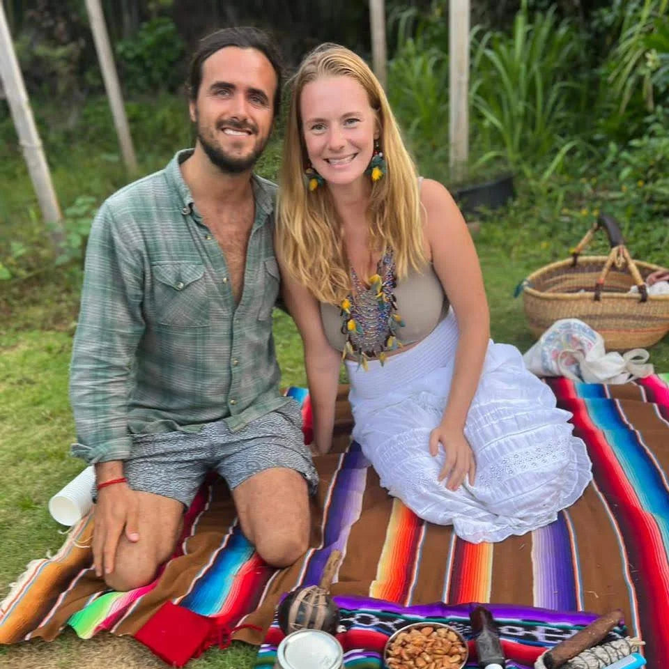 Two people sitting on a colorful blanket outdoors with cacao and tobacco and sage.