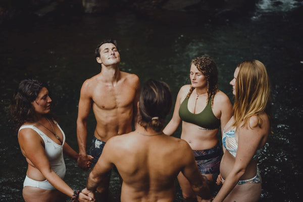 A group of 5 people holding hands in a circle standing in water with their eyes closed. 