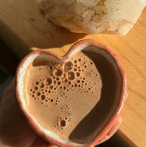 Heart-shaped ceramic mug filled with a frothy brown beverage on a wooden surface.